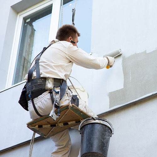Pintor com cinto de segurança pintando a parede externa de uma casa com rolo